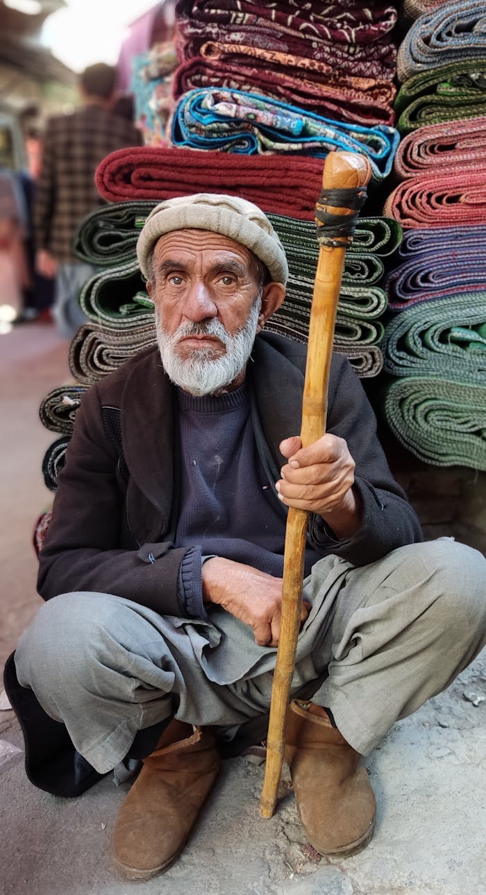 Portrait of an elderly man with a wooden stick seated beside colorful textiles at a market in Kabul, Afghanistan.