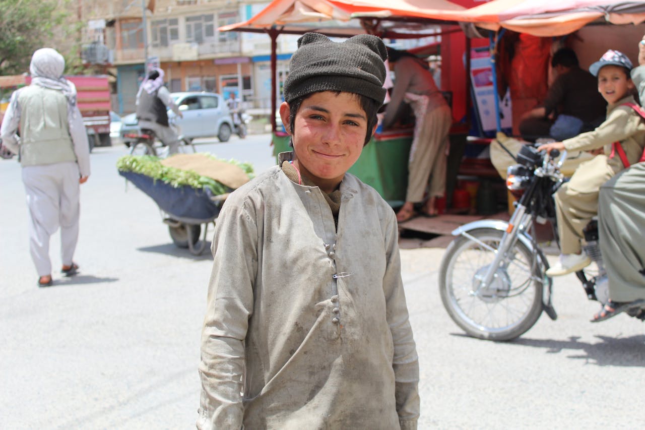 A young boy smiles in a bustling Ghazni street market with vibrant activity around.