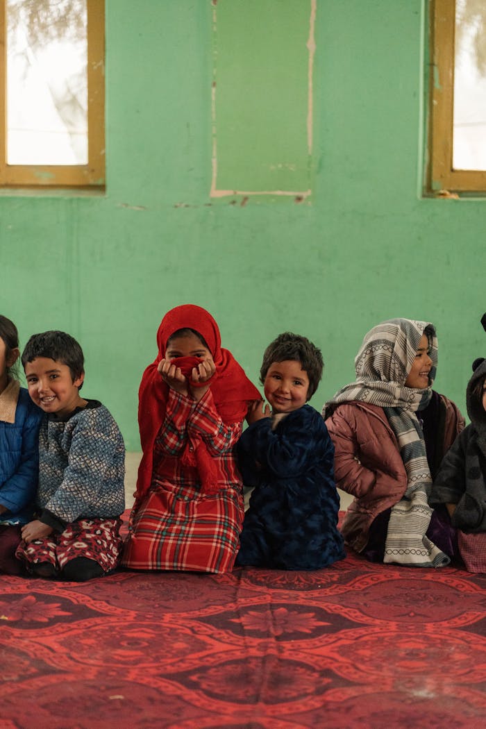 Group of Afghan children sitting together indoors, dressed warmly in colorful clothing, evoking a sense of community and culture.