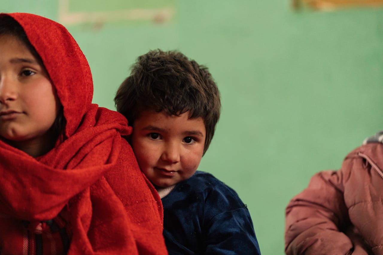 Two Afghan children in traditional clothing, captured in a candid moment indoors.